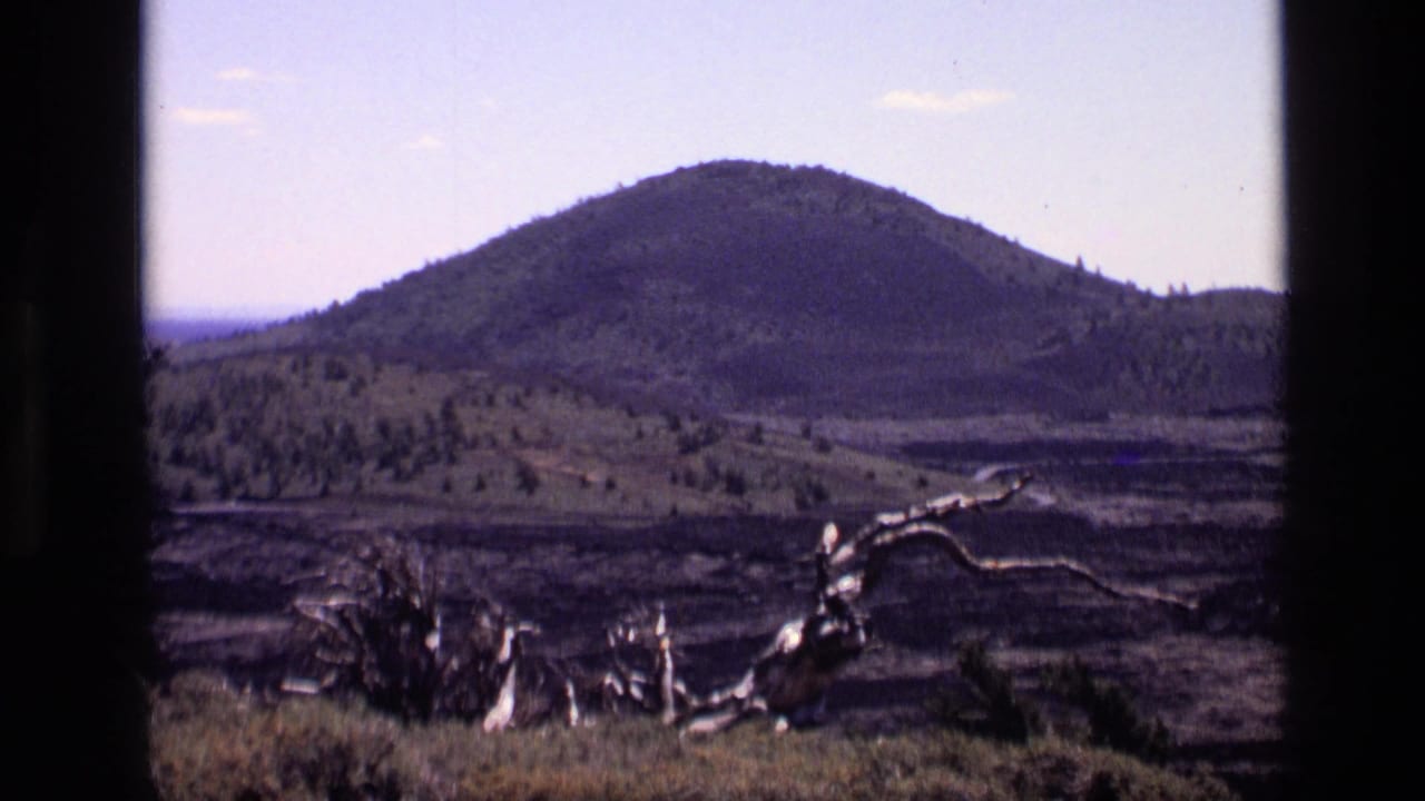 1972 Idaho 8mm frame showing a rounded mountain, sparse forest, dark terrain, and weathered dead wood in the foreground.