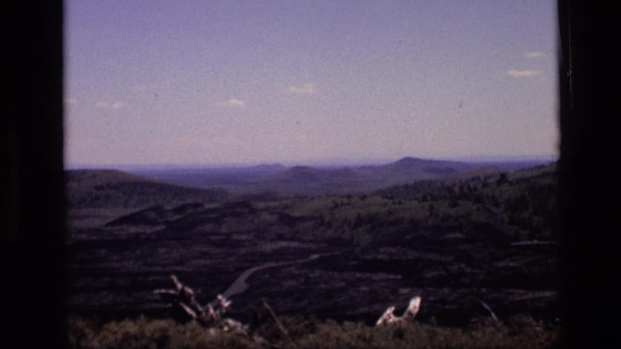 1972 Idaho 8mm frame showing a wide rural hillside view with distant ridges, brush in the foreground, and a winding road below.