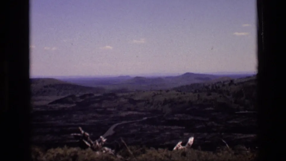 1972 Idaho 8mm frame showing a wide rural hillside view with distant ridges, brush in the foreground, and a winding road below.