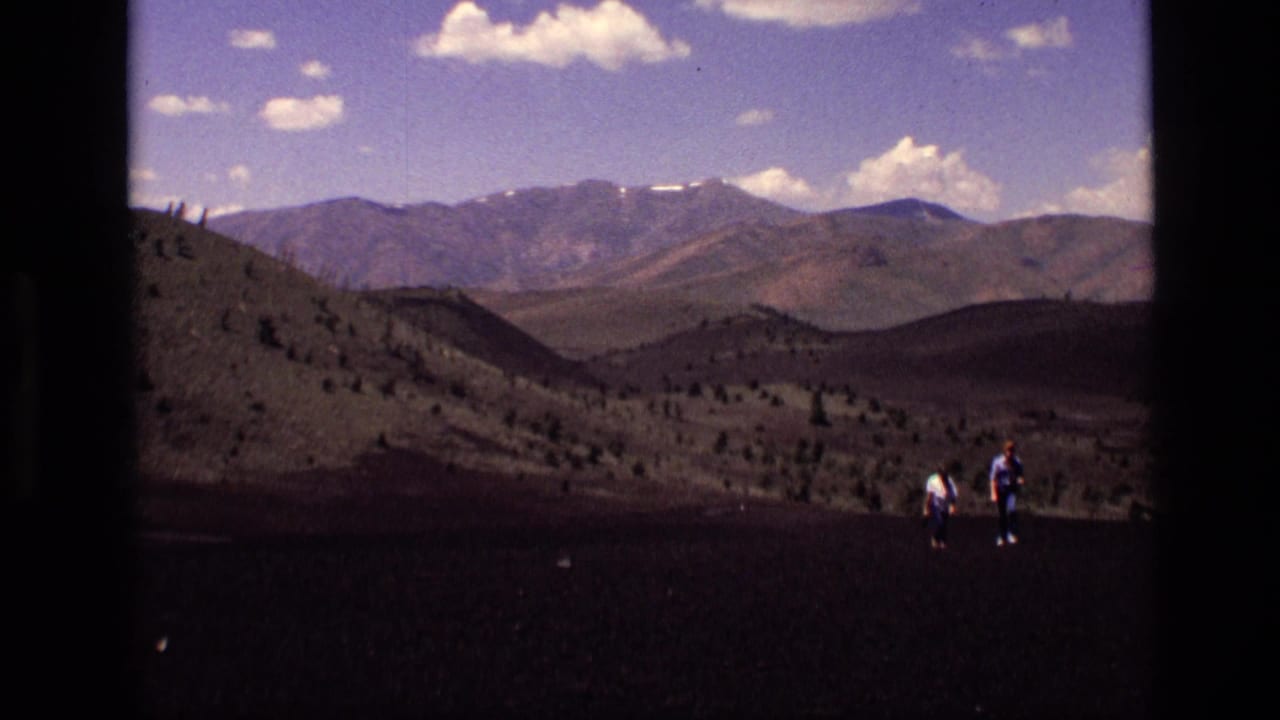 Two distant hikers cross dark volcanic-looking ground in Idaho, with mountain ridges and scattered clouds in the background.