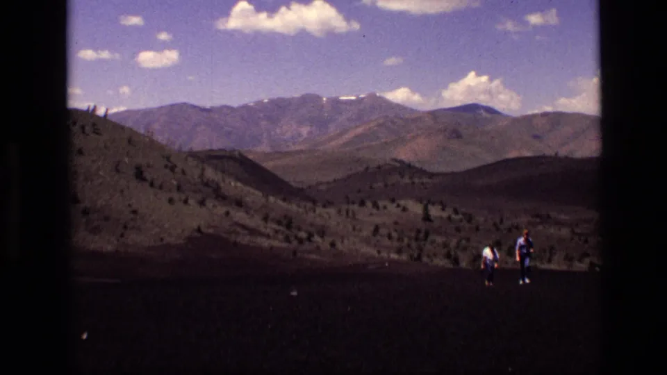 Two distant hikers cross dark volcanic-looking ground in Idaho, with mountain ridges and scattered clouds in the background.