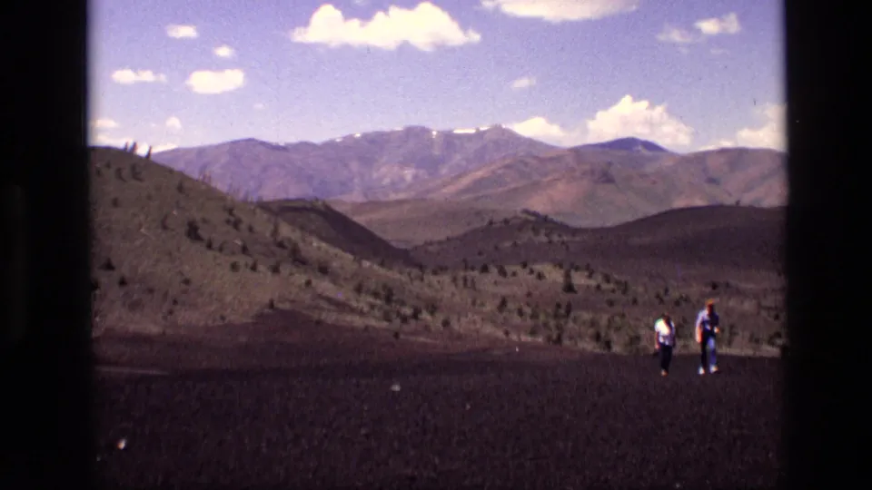 Distant hikers in casual 1970s attire appear against Idaho mountain views, dark terrain, and sparse vegetation.