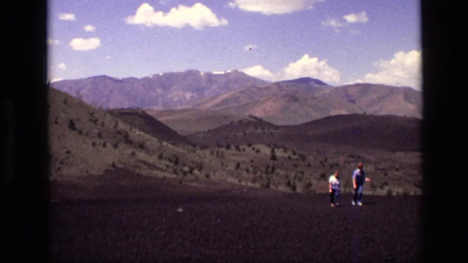 Two distant hikers appear on dark volcanic-looking Idaho terrain, with rolling hills and a broad mountain range beyond.