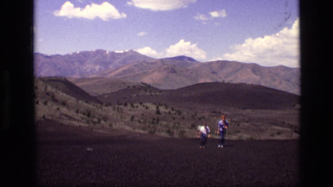 Two hikers stand on dark rocky ground with mountain ridges and clouds behind them.