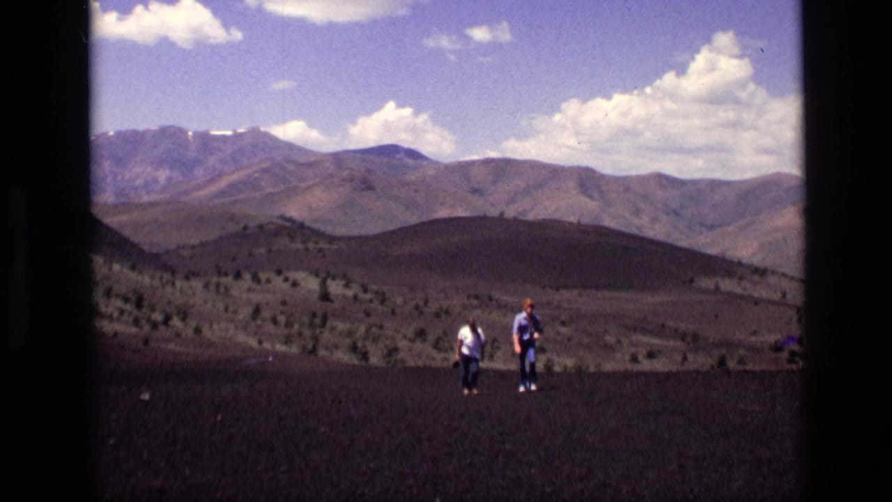 Two hikers cross a dark open slope with distant mountains under a cloudy blue sky.
