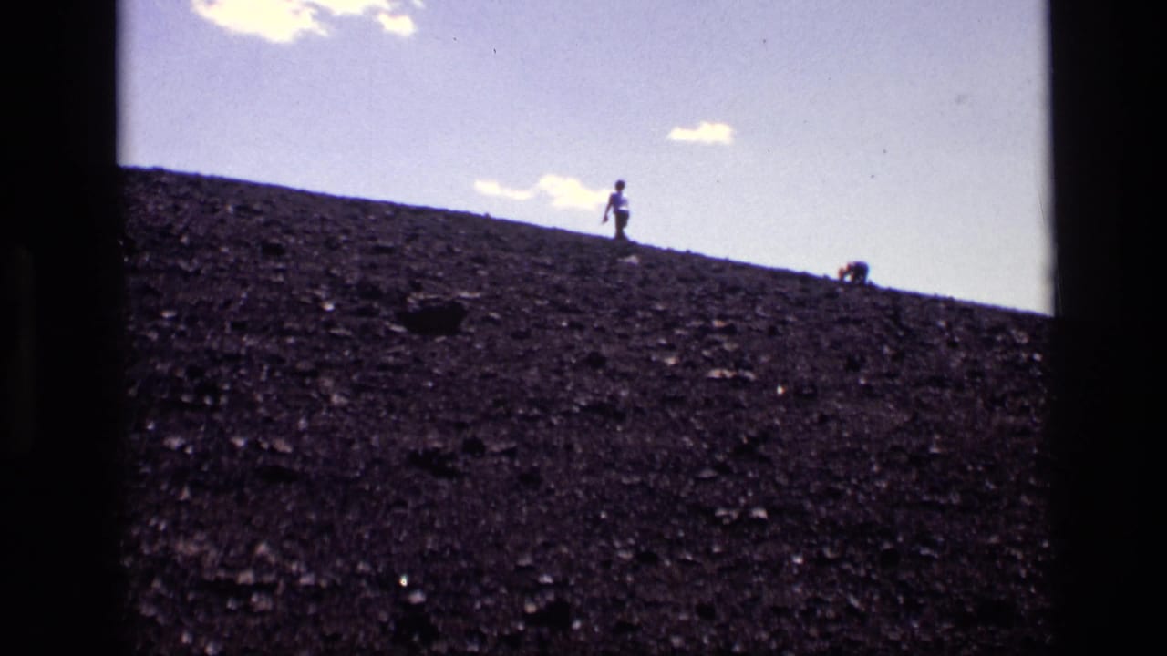 1972 Idaho 8mm frame showing a distant walker silhouetted on a dark volcanic hillside under pale sky and small clouds.