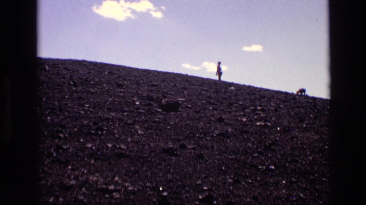 1972 Idaho home-movie frame with a small visitor figure on a dark cinder-like hillside and sparse clouds overhead.