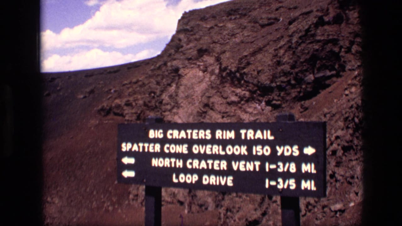 1972 Idaho 8mm frame showing a Big Craters Rim Trail directional sign against rocky volcanic terrain and clouds.