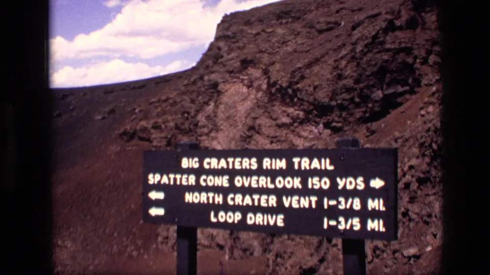 1972 Idaho 8mm frame showing a Big Craters Rim Trail directional sign against rocky volcanic terrain and clouds.