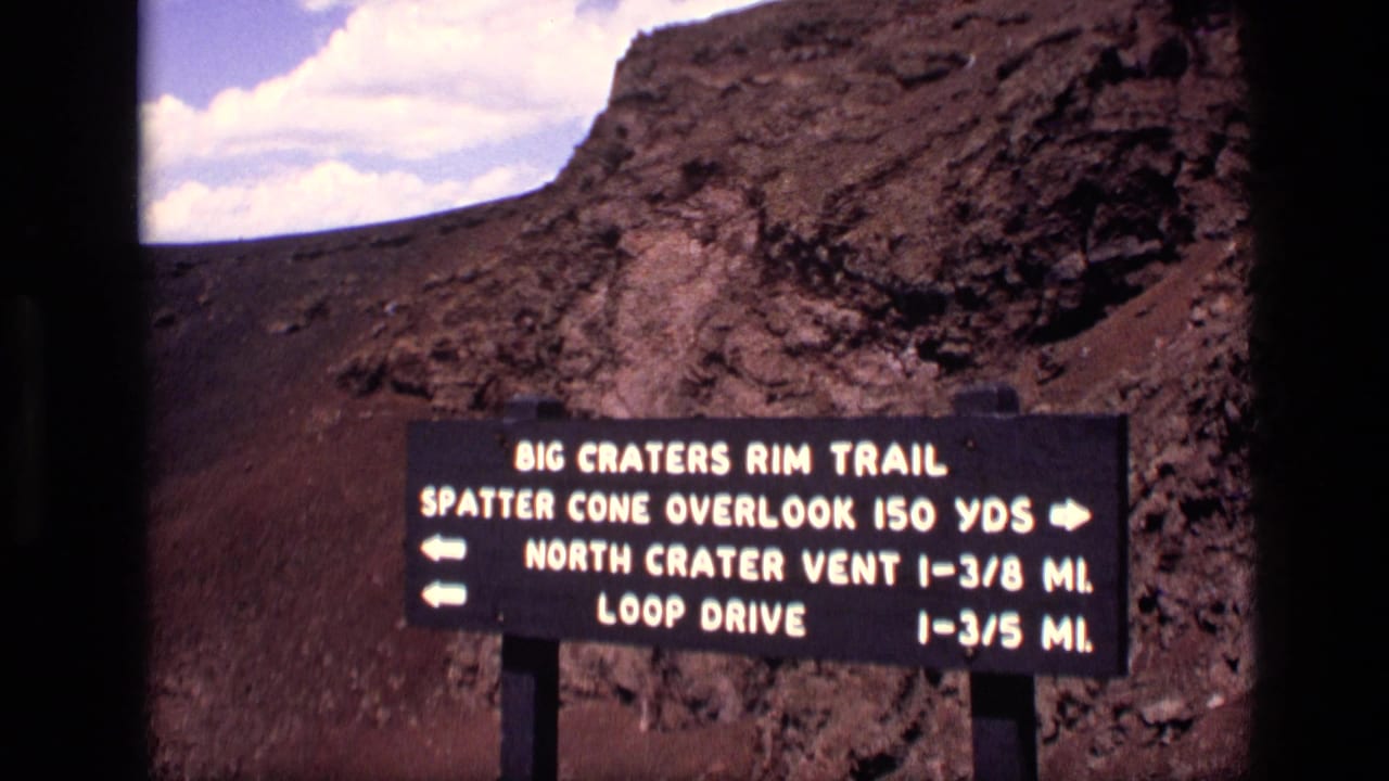1972 Idaho home-movie still with the Big Craters Rim Trail sign framed before rugged volcanic rock and open sky.