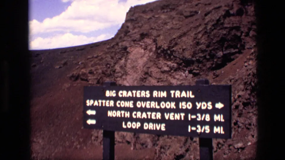 1972 Idaho home-movie still with the Big Craters Rim Trail sign framed before rugged volcanic rock and open sky.