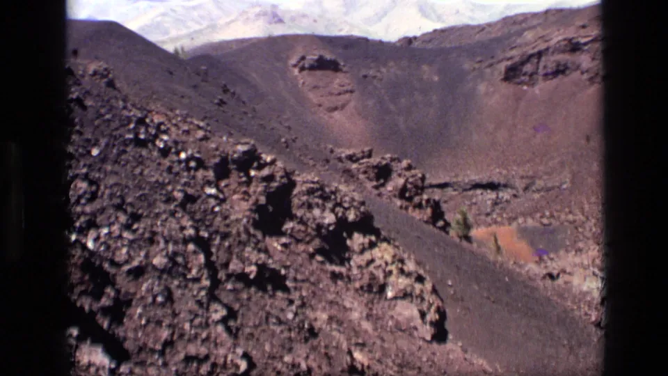1972 Idaho home-movie frame showing a broad volcanic crater landscape with rocky foreground and pale clouds above.