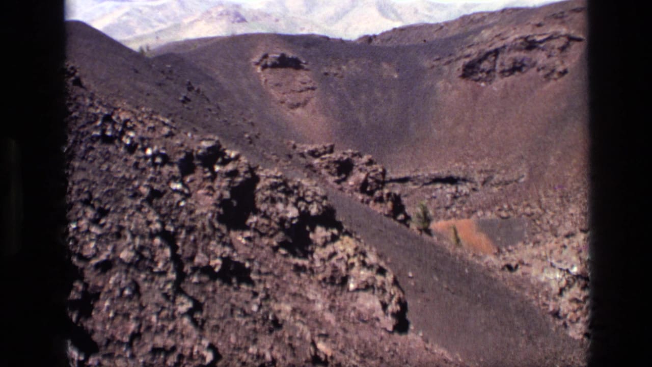 Dark volcanic crater slopes with rocky foreground and pale mountains in the distance.