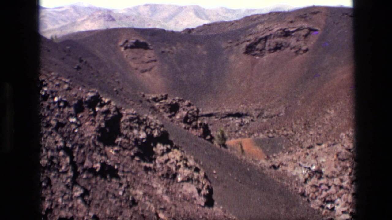 Rugged black and brown crater wall with lava rocks and distant mountains.