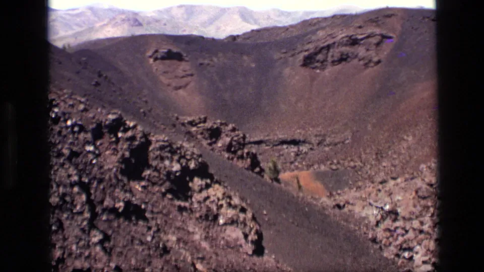 1972 Idaho home-movie frame of a volcanic crater wall, dark rocky outcrops, scattered boulders, and mountains along the horizon.