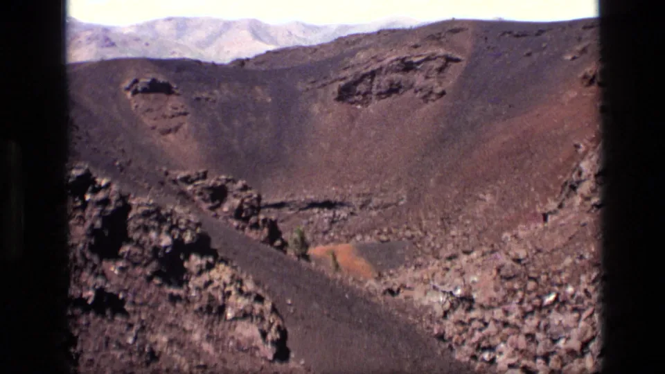 1972 Idaho 8mm frame showing a broad brown volcanic basin, black lava rock, scattered stones, and pale mountains in the background.