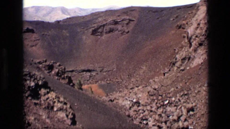 1972 Idaho home-movie frame with dark lava rocks at the edges, a brown crater slope, loose boulders, and a pale mountain range beyond.