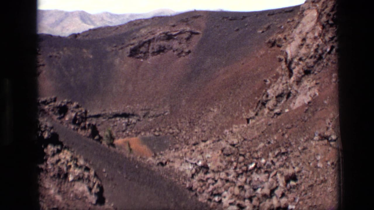 1972 Idaho 8mm frame showing dark volcanic crater walls, reddish-brown cinder ground, rough rock, and distant ridges.