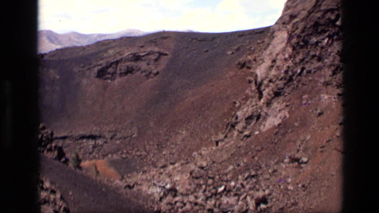 1972 Idaho home-movie view of a scorched volcanic slope, rocky outcrop, crater wall, and pale clouded sky.