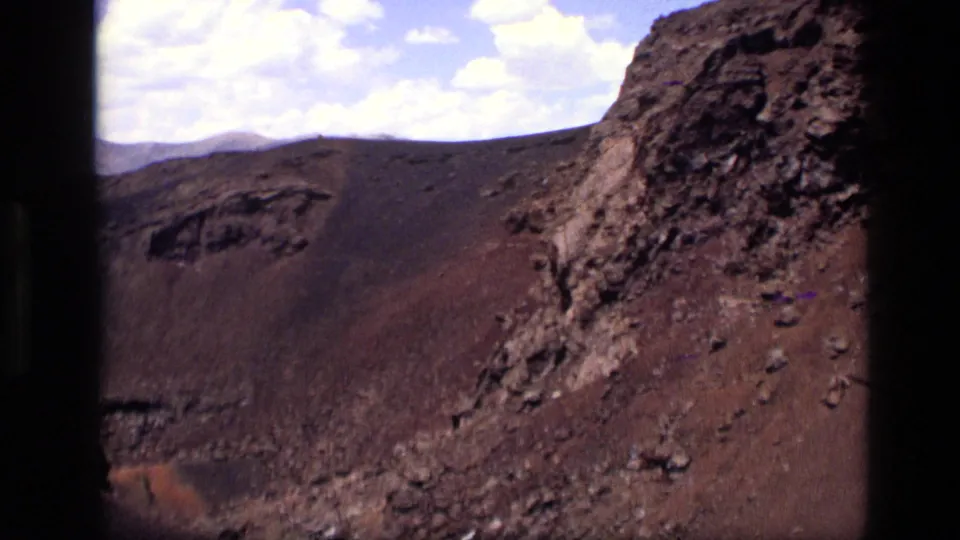 1972 Idaho 8mm still of barren volcanic hillside, dark crater rim, reddish cinder earth, and scattered white clouds.