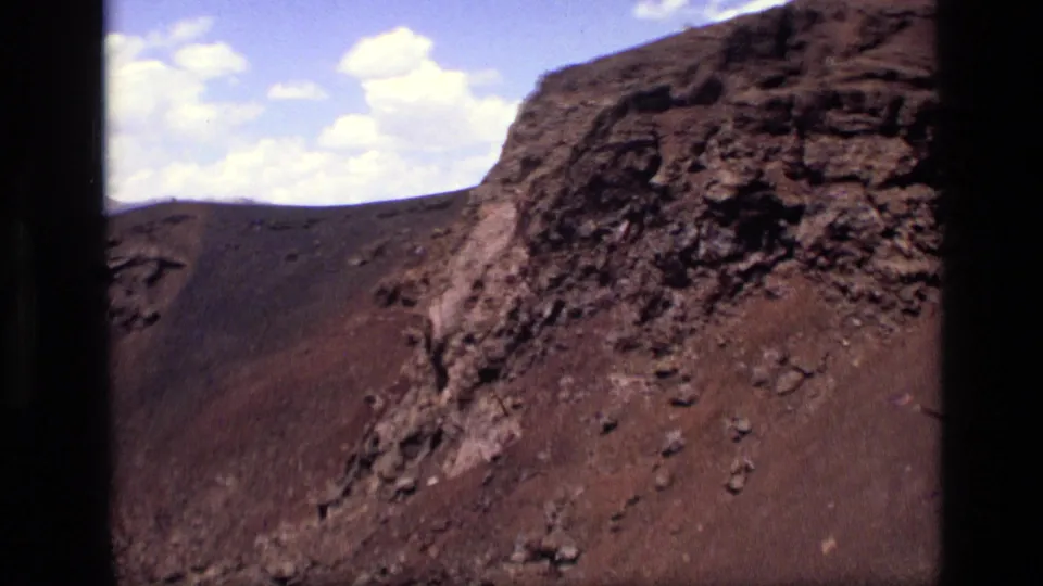 1972 Idaho 8mm frame with jagged lava rock, dark volcanic slopes, scattered clouds, and visible film-frame borders.