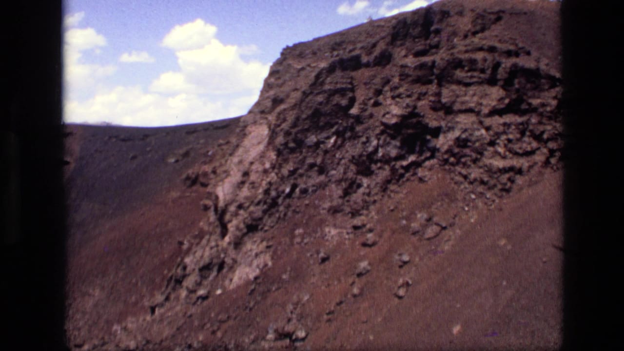Dark volcanic cliff and brown rocky slope beneath a blue sky with small clouds.