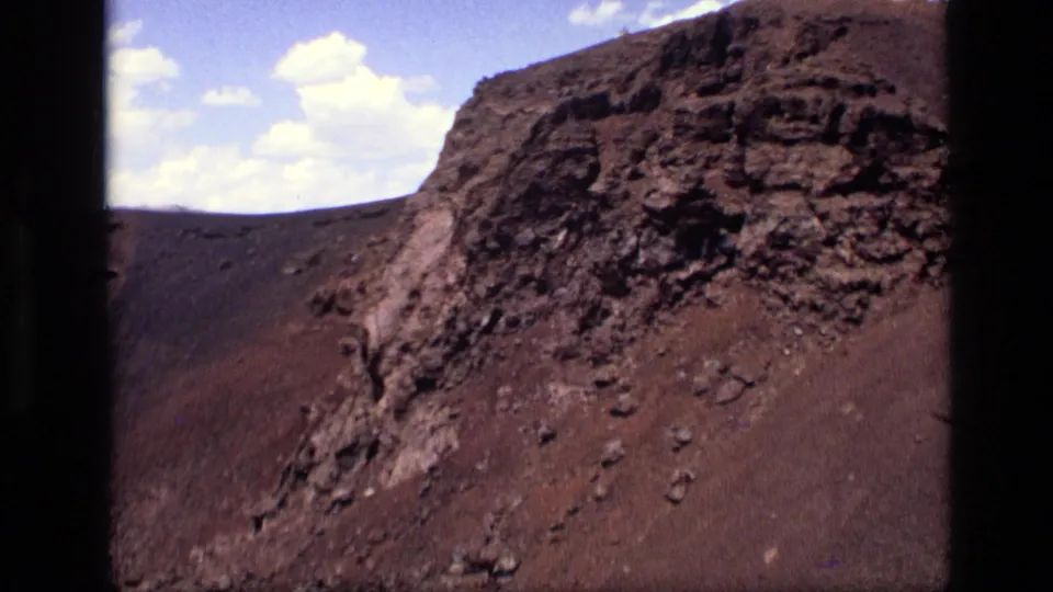 1972 Idaho 8mm frame showing a rugged dark volcanic rock face and steep brown slope under scattered clouds.