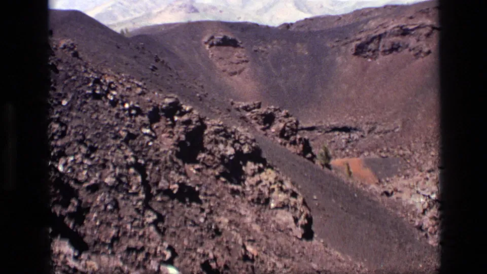 1972 Idaho 8mm frame showing a wide barren volcanic basin, dark foreground rocks, and layered ridges in the distance.