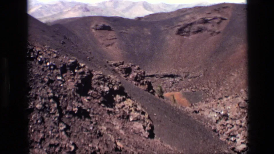 1972 Idaho 8mm frame looking across a barren volcanic basin toward dark slopes and pale mountains beyond.