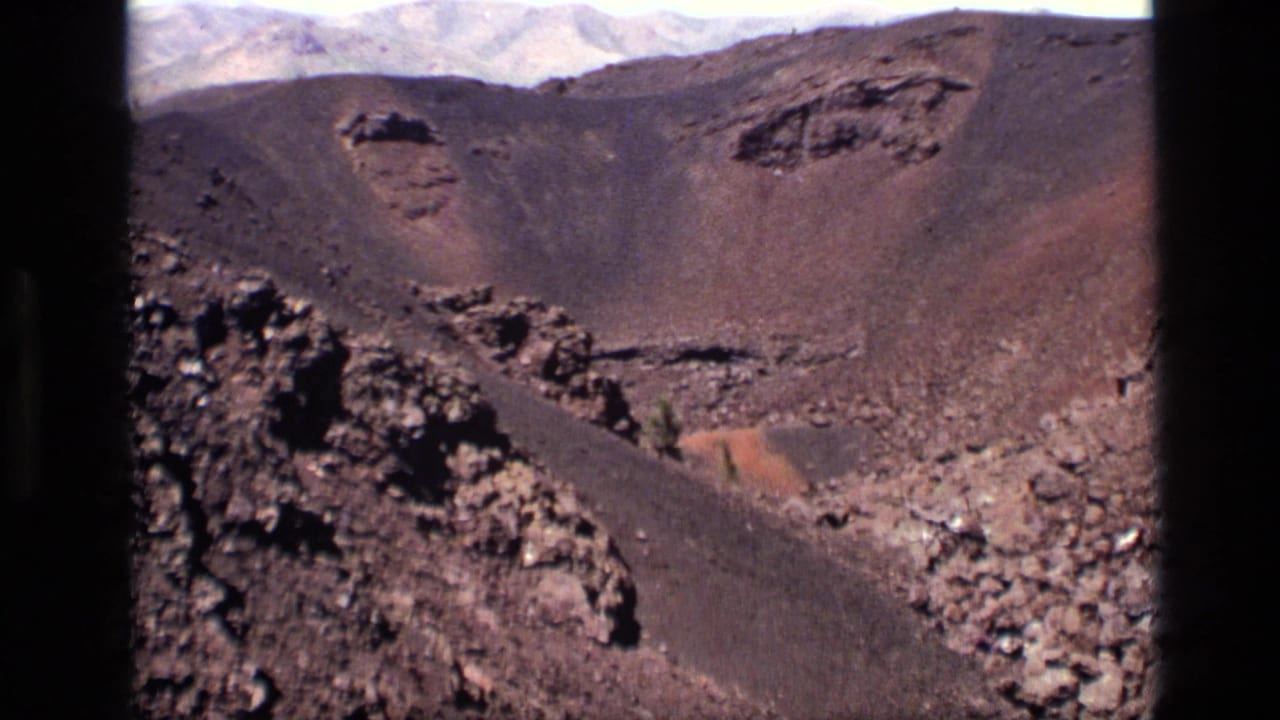 1972 Idaho 8mm frame showing a rocky volcanic crater slope, dark lava rubble, reddish ground, and distant mountains under a bright sky.