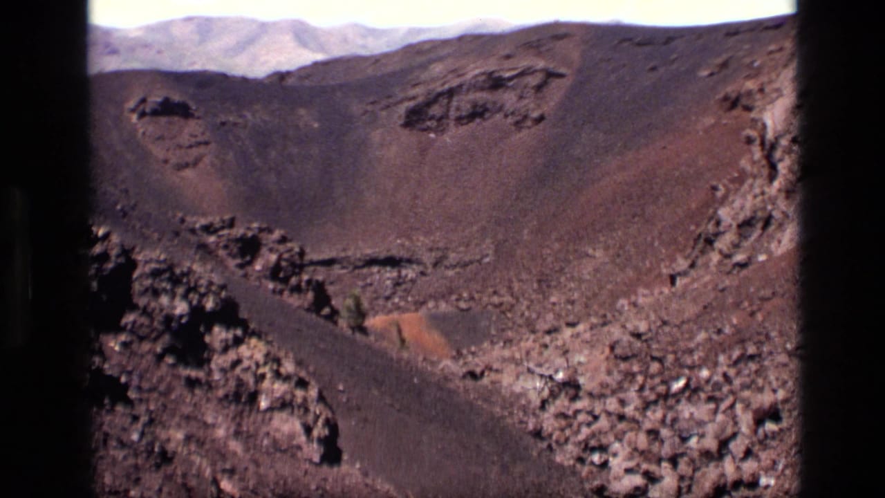 1972 Idaho home-movie frame looking across a dark volcanic crater interior with rough lava rock, reddish gravel, and pale mountains in the distance.