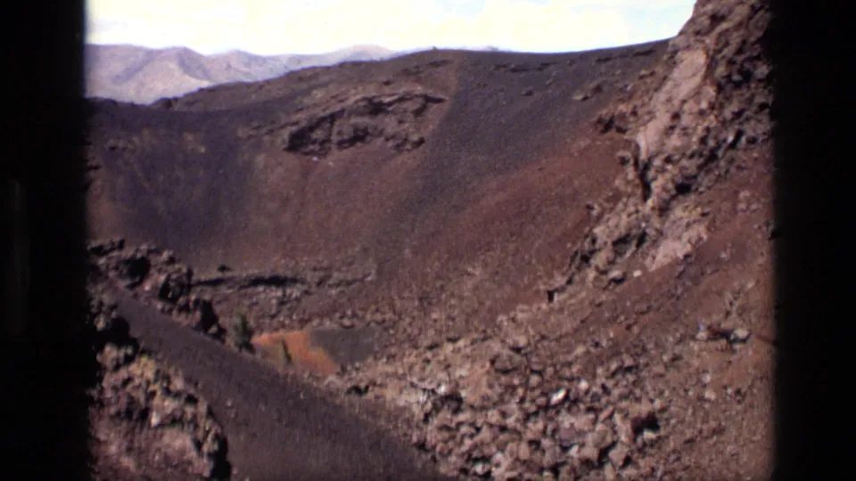 1972 Idaho 8mm frame showing the pan moving along a rocky crater edge, with dark slopes, loose lava stones, and a reddish patch below.