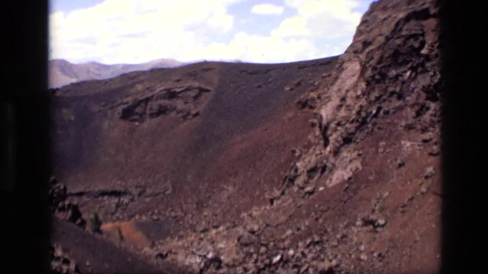 1972 Idaho home-movie frame ending on a closer rocky crater wall, dark volcanic slope, scattered stones, and bright clouded sky.