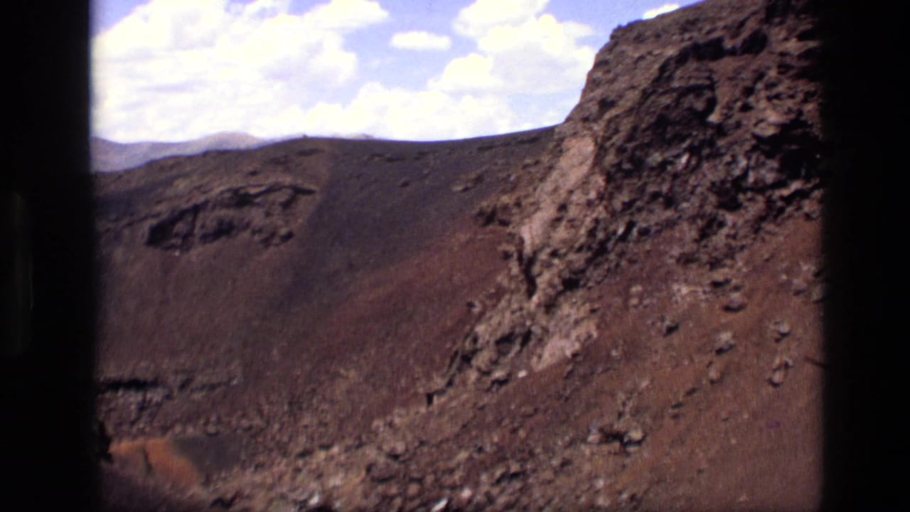 1972 Idaho 8mm frame showing a barren red-brown slope, dark volcanic-looking rock, loose scree, and scattered clouds overhead.