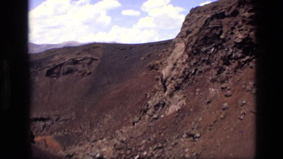 1972 Idaho 8mm frame showing a barren red-brown slope, dark volcanic-looking rock, loose scree, and scattered clouds overhead.