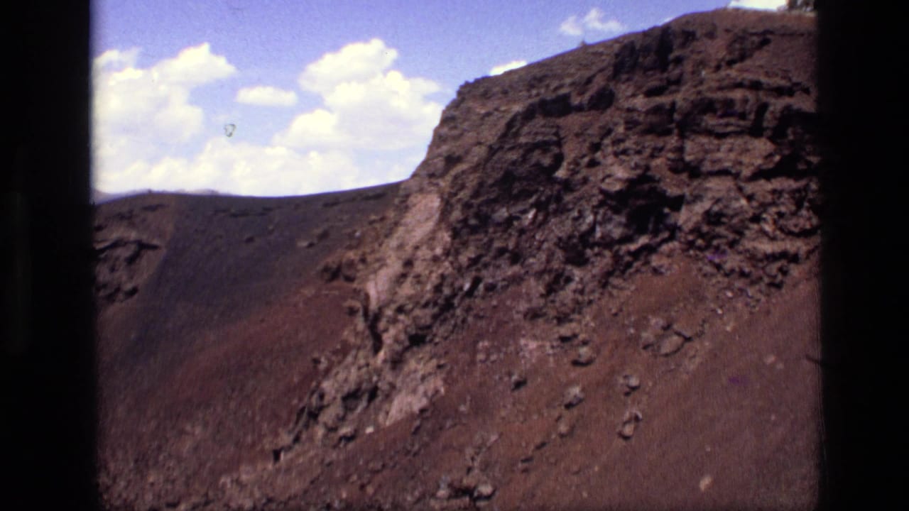 1972 Idaho home-movie frame of a rocky red-brown hillside with a prominent dark outcrop and sparse cloud cover in the sky.