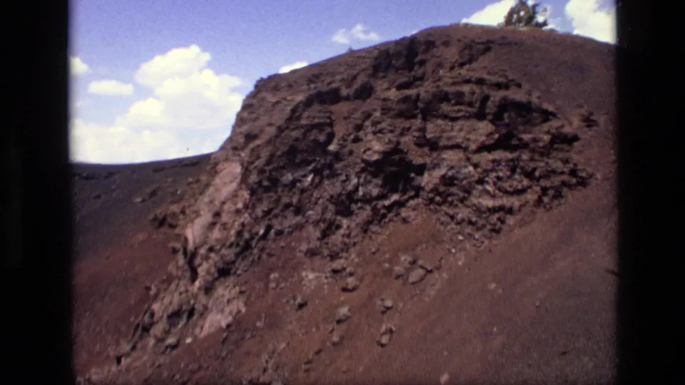 1972 Idaho 8mm frame with a closer view of rough red-brown volcanic-looking rock, scattered stones, and a small strip of clouded sky.
