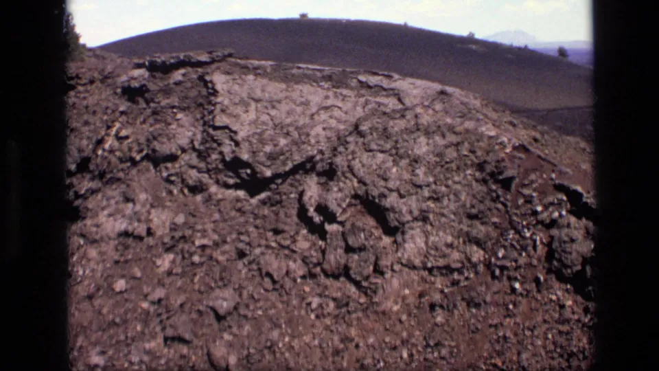 1972 Idaho 8mm frame looking over rough dark rock toward a rounded, vegetation-free hill beneath a pale clouded sky.