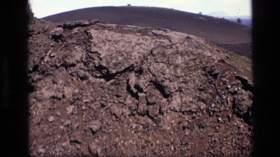 1972 Idaho 8mm frame showing broken earth and dark rocks in front of a distant rounded hill under a pale sky.