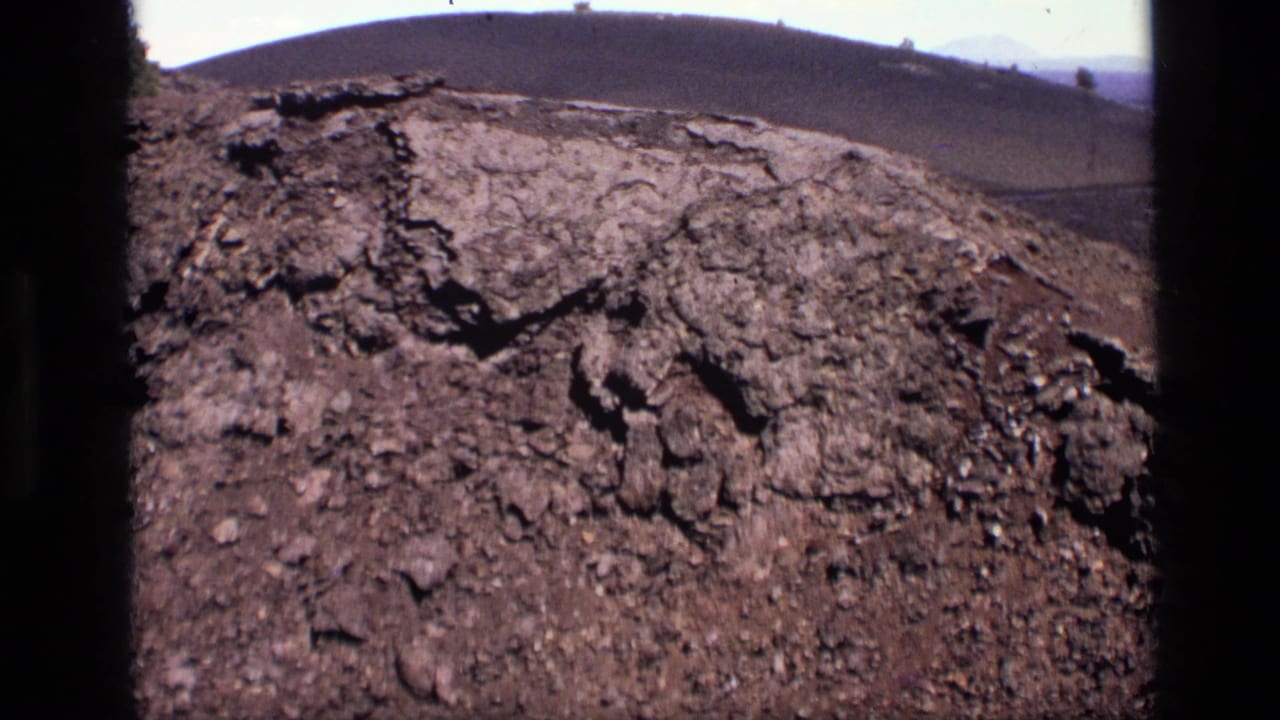 1972 Idaho 8mm frame tightening on jagged cracked dirt and weathered rocks, with a soft hill line in the distance.
