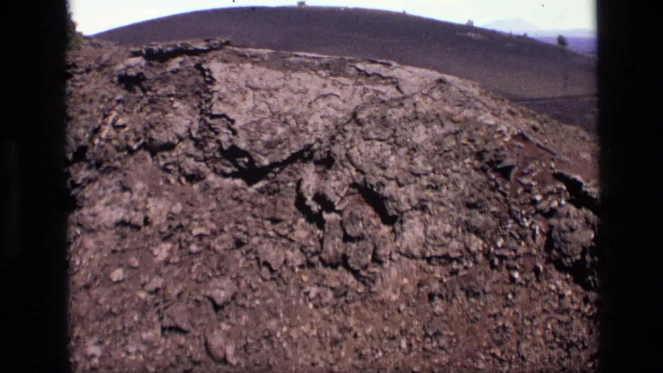 1972 Idaho 8mm frame tightening on jagged cracked dirt and weathered rocks, with a soft hill line in the distance.