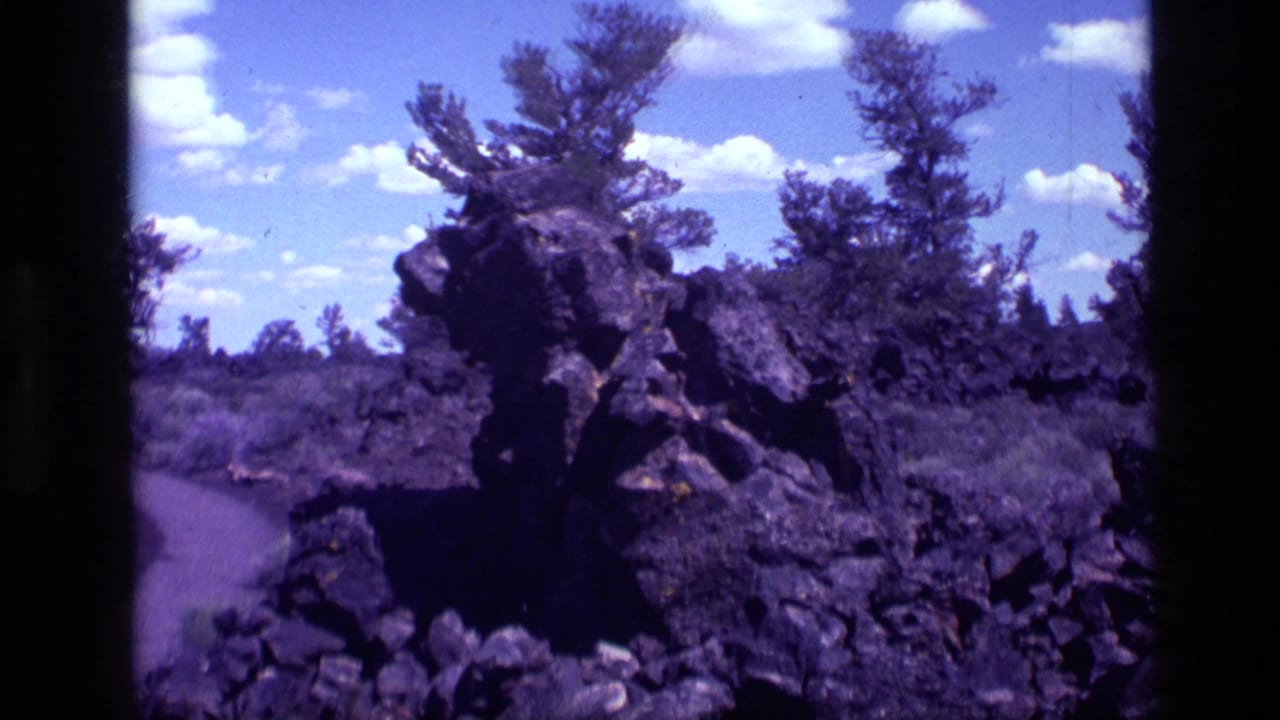 Dark roadside rock formation with pine trees, scrub, blue sky, and film-frame borders.