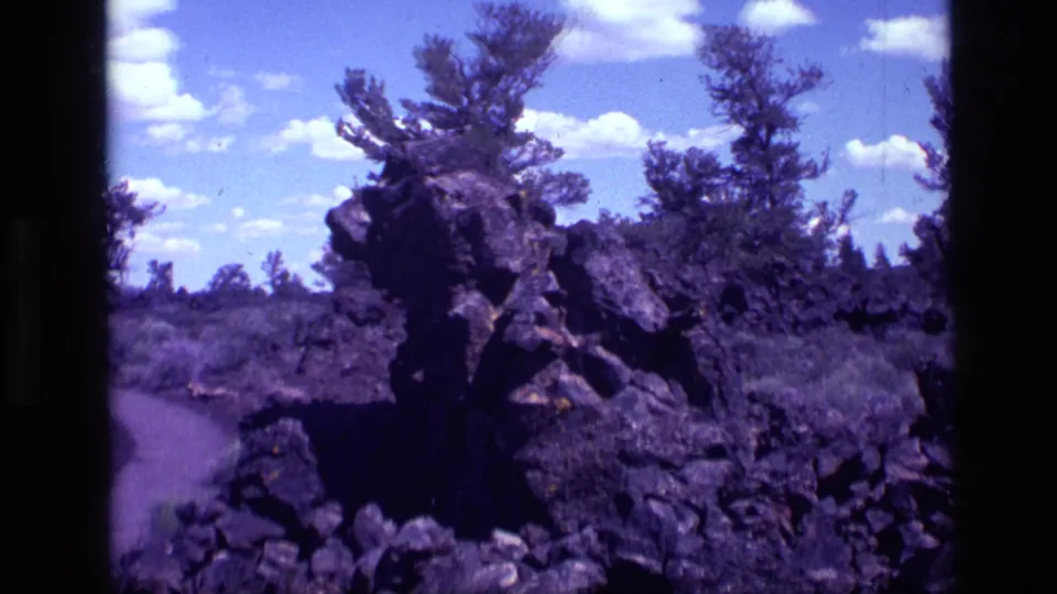 1972 Idaho 8mm frame showing a rugged dark rock outcrop beside vegetation, with pines and scattered clouds above.