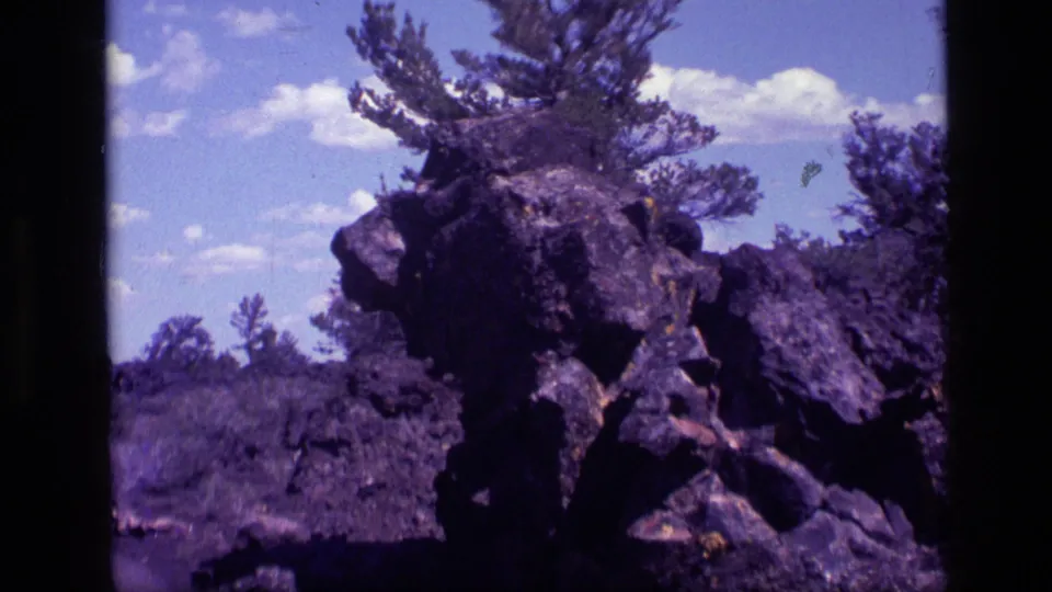 Closer 1972 Idaho 8mm view of the dark roadside rock, with a pine rising behind it and brushland beyond.