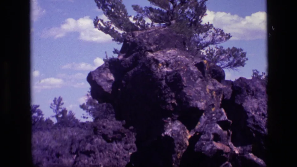 1972 Idaho 8mm frame showing the roadside rock formation in closer framing, with pines and scattered clouds overhead.
