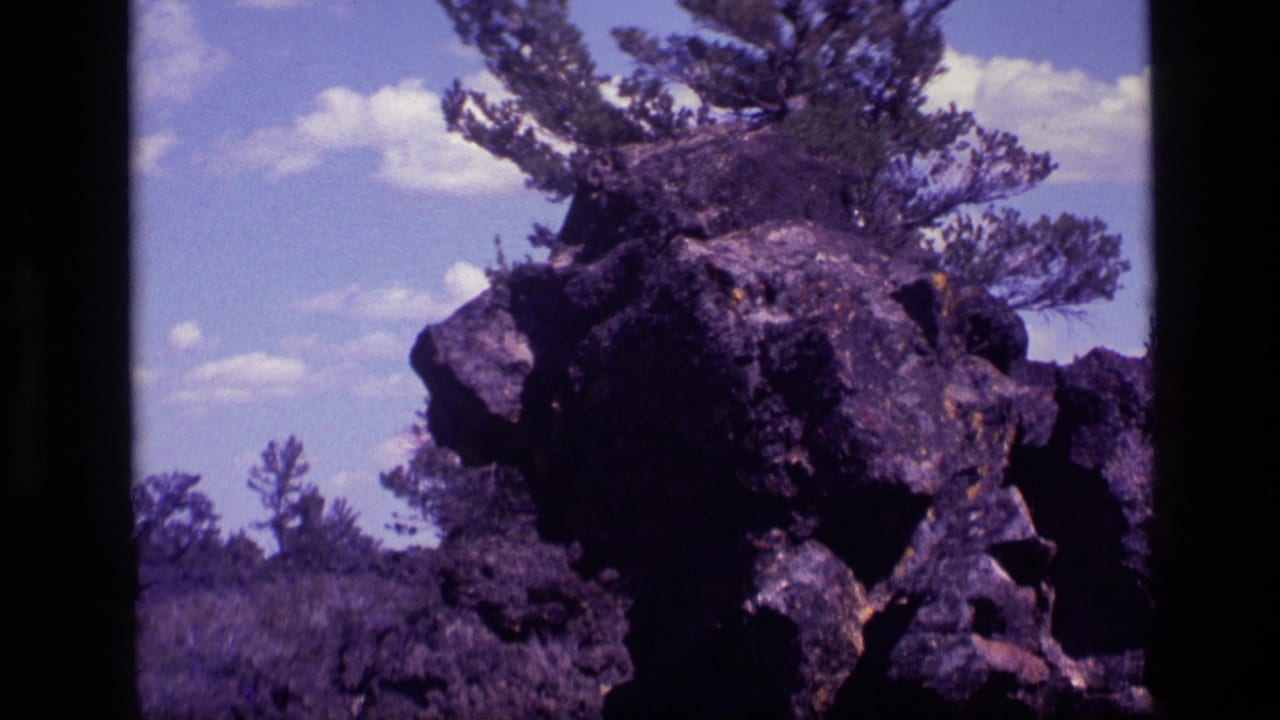 1972 Idaho 8mm frame showing a dark rock outcrop among brush and pine branches under a blue sky with scattered clouds.