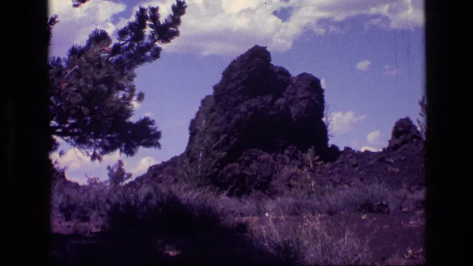1972 Idaho 8mm view of a jagged dark rock formation in dry brush with pine trees and a bright clouded sky.