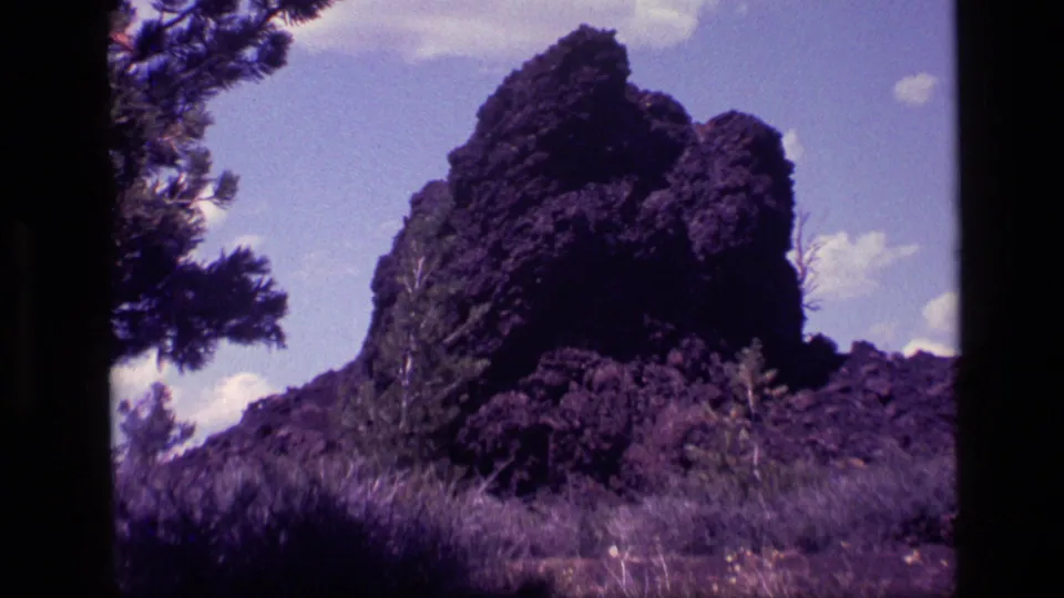 1972 Idaho home-movie frame of a tall dark rock formation viewed across dry grass and brush, with a pine branch at left.