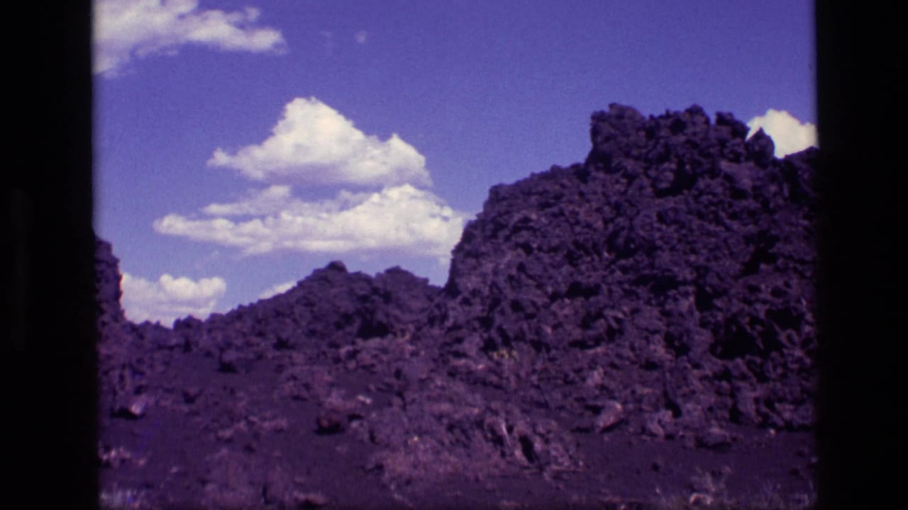 1972 Idaho 8mm frame with rough volcanic-looking rock piles, broken stones, and a bright sky with scattered white clouds.