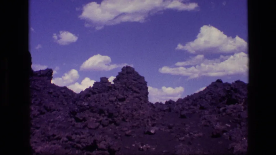 1972 Idaho 8mm frame showing a dark rocky field, jagged mound-like formations, and scattered clouds in a saturated blue sky.